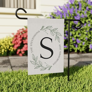 May include: A personalized garden flag with the name "The Stevensons" and the letter "S" in a circular design with leaves. The flag is cream-colored and reads "Established 2026". The flag is displayed on a black metal stand in a garden setting.