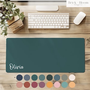 Op de afbeelding: Teal bureaumat met de naam "Olivia" in witte letters. De mat is rechthoekig en ligt op een houten bureau, naast een toetsenbord, muis en een kleine plant. Een kleurenpalet van verschillende tinten wordt eronder weergegeven.