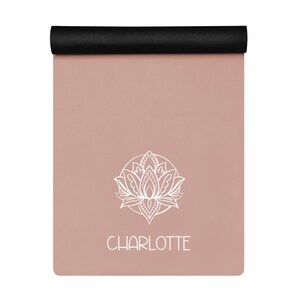 May include: A rolled-up yoga mat with a black top and a dusty rose surface. A white lotus flower design is centered on the mat, with the name "CHARLOTTE" printed below it in white. The mat is designed for yoga and fitness, measuring approximately 173 cm in length.