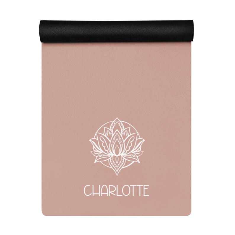 May include: A rolled-up yoga mat with a black top and a dusty rose surface. A white lotus flower design is centered on the mat, with the name "CHARLOTTE" printed below it in white. The mat is designed for yoga and fitness, measuring approximately 173 cm in length.