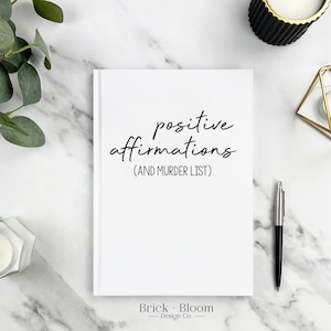 May include: White notebook with the text "positive affirmations (AND MURDER LIST)" on a marble surface with candles and greenery.