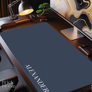 May include: A navy blue desk mat with the name "ALEXANDER" printed in white. The mat is on a wooden desk with a lamp, bonsai tree, and computer monitor. The desk mat is from Brick + Bloom Design Co.