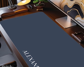 May include: A large, rectangular, dark navy blue desk mat featuring the name ALEXANDER printed in white capital letters along the bottom left corner. The mat has a smooth, solid-colored surface and is positioned on a dark wooden desk.