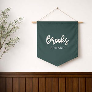May include: A dark green pennant banner with the name "Brooks" in a white script font above the name "EDWARD" in a white sans-serif font. The banner is suspended from a wooden dowel with a natural twine hanger.