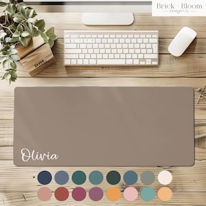 May include: A taupe desk mat with the name "Olivia" in white script. The mat is rectangular and sits on a wooden desk, with a keyboard, mouse, and plant visible. A color palette of 12 colors is displayed below the mat.