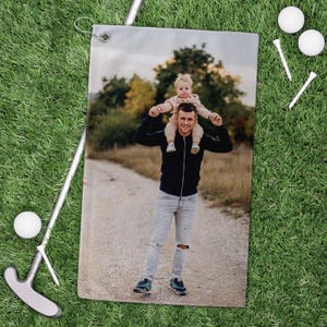 May include: Golf towel featuring a photograph of a man carrying a child on his shoulders. The towel is rectangular with a metal grommet for hanging. Golf balls, tees, and a putter are on the green grass.