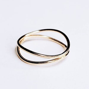 Puede incluir: Un anillo de tono dorado con un diseño simple y minimalista. El anillo presenta dos bandas que se cruzan entre sí, creando un aspecto delicado y moderno.