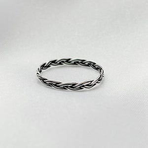 May include: A silver ring featuring a braided pattern. The circular ring is crafted from metal and is set against a white backdrop. The design gives the ring a textured appearance.