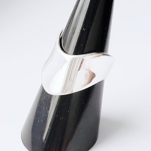 Könnte beinhalten: Ein polierter Silberring mit einem glatten, asymmetrischen Design. Der Ring wird auf einem schwarzen, kegelförmigen Ständer vor einem weißen Hintergrund präsentiert. Die Oberfläche des Rings reflektiert das Licht und unterstreicht sein schlankes, modernes Aussehen.