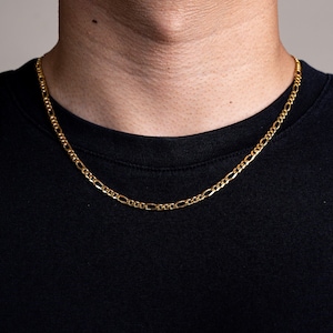 Pode incluir: Um colar de corrente de ouro com um design clássico figaro. A corrente é usada por uma pessoa com uma camisa preta.