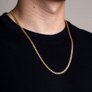 May include: A gold chain necklace with a curb link style. The chain is worn by a person with a black shirt.