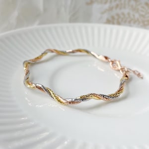 Könnte beinhalten: Ein Armband aus einer verdrehten Kette in Gold, Silber und Roségold. Das Armband liegt auf einem weißen Teller.