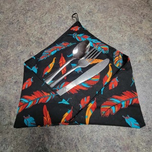 May include: A black fabric napkin with a colourful feather pattern. The napkin is folded in half and has a silver fork, knife, and spoon inside.