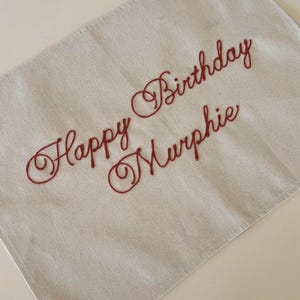 May include: Cream-colored banner with the embroidered text "Happy Birthday Murphie" in a cursive, reddish-brown font. The banner has a reinforced corner with a metal grommet.