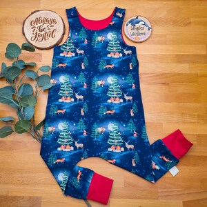 Op de afbeelding: Een blauwe en rode kerst-baby romper met een bosdierprint. De romper heeft verschillende dieren, waaronder herten, vossen en uilen, evenals kerstbomen en sterren. De romper heeft een rode rand rond de hals en armsgaten. De tekst "Always be Joyful" is zichtbaar op een houten bord op de achtergrond.