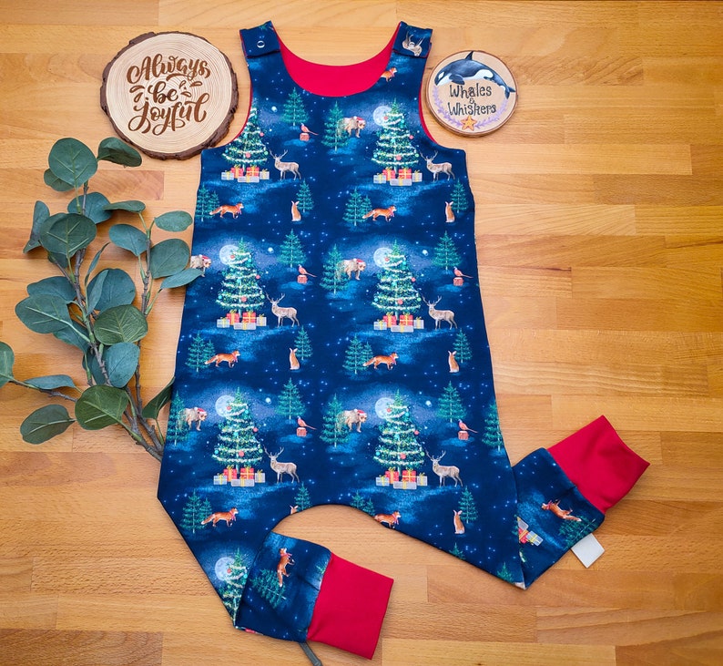 Op de afbeelding: Een blauwe en rode kerst-baby romper met een bosdierprint. De romper heeft verschillende dieren, waaronder herten, vossen en uilen, evenals kerstbomen en sterren. De romper heeft een rode rand rond de hals en armsgaten. De tekst "Always be Joyful" is zichtbaar op een houten bord op de achtergrond.