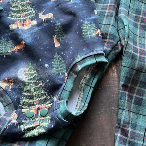 Op de afbeelding: Een blauwe en groene geruite stof met een kerstthema. De print heeft een donkerblauwe achtergrond met een witte maan, sterren en een verscheidenheid aan bosdieren, waaronder herten, vossen en konijnen. De stof heeft ook kerstbomen die versierd zijn met ornamenten en cadeaus. De stof is perfect voor knutselprojecten, zoals kleding, dekens en kussens.
