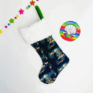 Op de afbeelding: Een donkerblauwe kerstsok met een witte nep bontkraag. De sok heeft een herhalend patroon van groene bomen, herten en vossen op een donkerblauwe achtergrond. De sok heeft een groene lus aan de bovenkant. De sok ligt op een witte achtergrond.