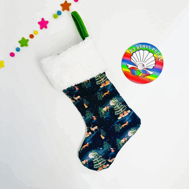 Op de afbeelding: Een donkerblauwe kerstsok met een witte nep bontkraag. De sok heeft een herhalend patroon van groene bomen, herten en vossen op een donkerblauwe achtergrond. De sok heeft een groene lus aan de bovenkant. De sok ligt op een witte achtergrond.