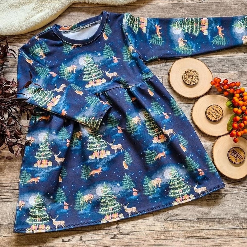 Op de afbeelding: Een blauwe, lange mouwen jurk met een feestelijke kerstboom en bosdier print. De jurk heeft een geplooide rok en een ronde halslijn.
