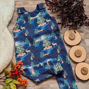 Op de afbeelding: Een donkerblauwe baby romper met een kerstboom- en bosdierprint. De romper heeft bandjes en drukknopen op de schouders. De romper ligt op een houten oppervlak met drie houten cirkels met de woorden "Nice", "Santa's coming" en "Naughty" erop.