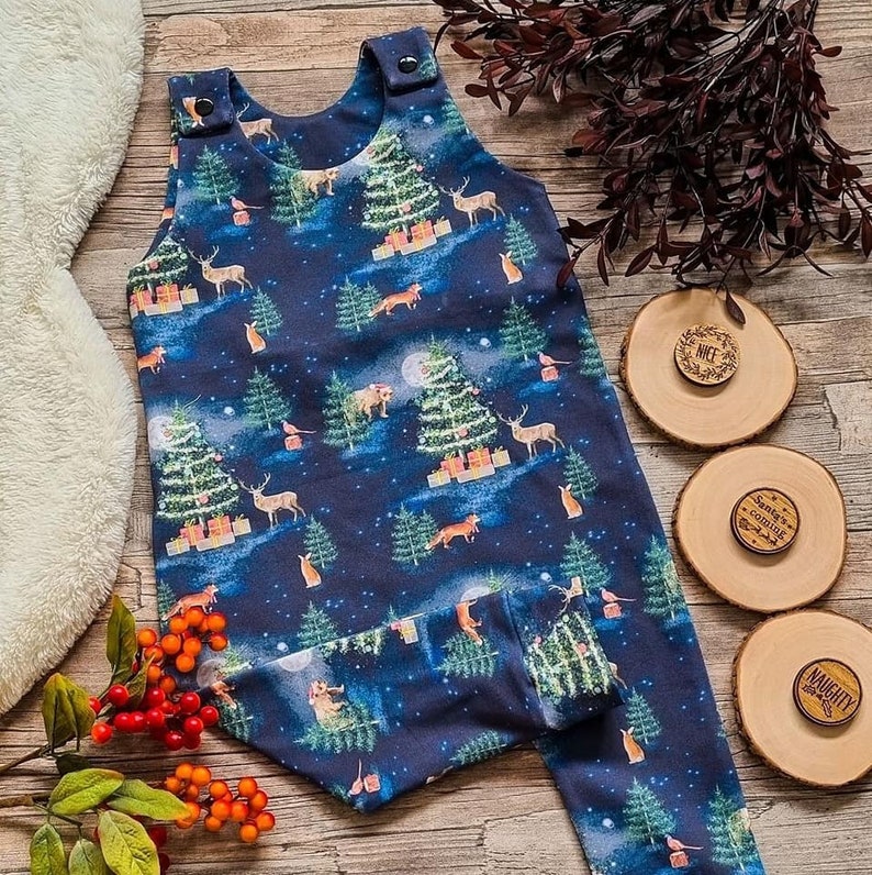Op de afbeelding: Een donkerblauwe baby romper met een kerstboom- en bosdierprint. De romper heeft bandjes en drukknopen op de schouders. De romper ligt op een houten oppervlak met drie houten cirkels met de woorden "Nice", "Santa's coming" en "Naughty" erop.