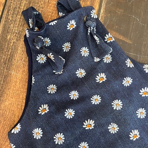 May include: A dark blue denim overall with a white daisy print. The straps have a knotted detail and a brown button.