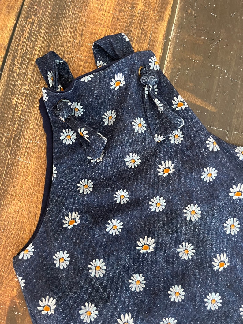 May include: A dark blue denim overall with a white daisy print. The straps have a knotted detail and a brown button.