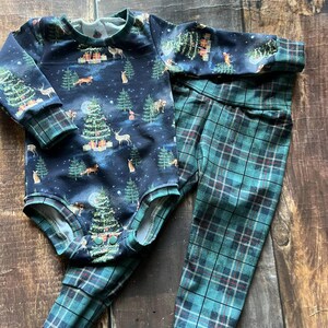 Op de afbeelding: Een baby-outfitset met een longsleeve bodysuit met een donkerblauwe achtergrond en een feestelijke kerstboomprint. De broek is groen en zwart geruit.