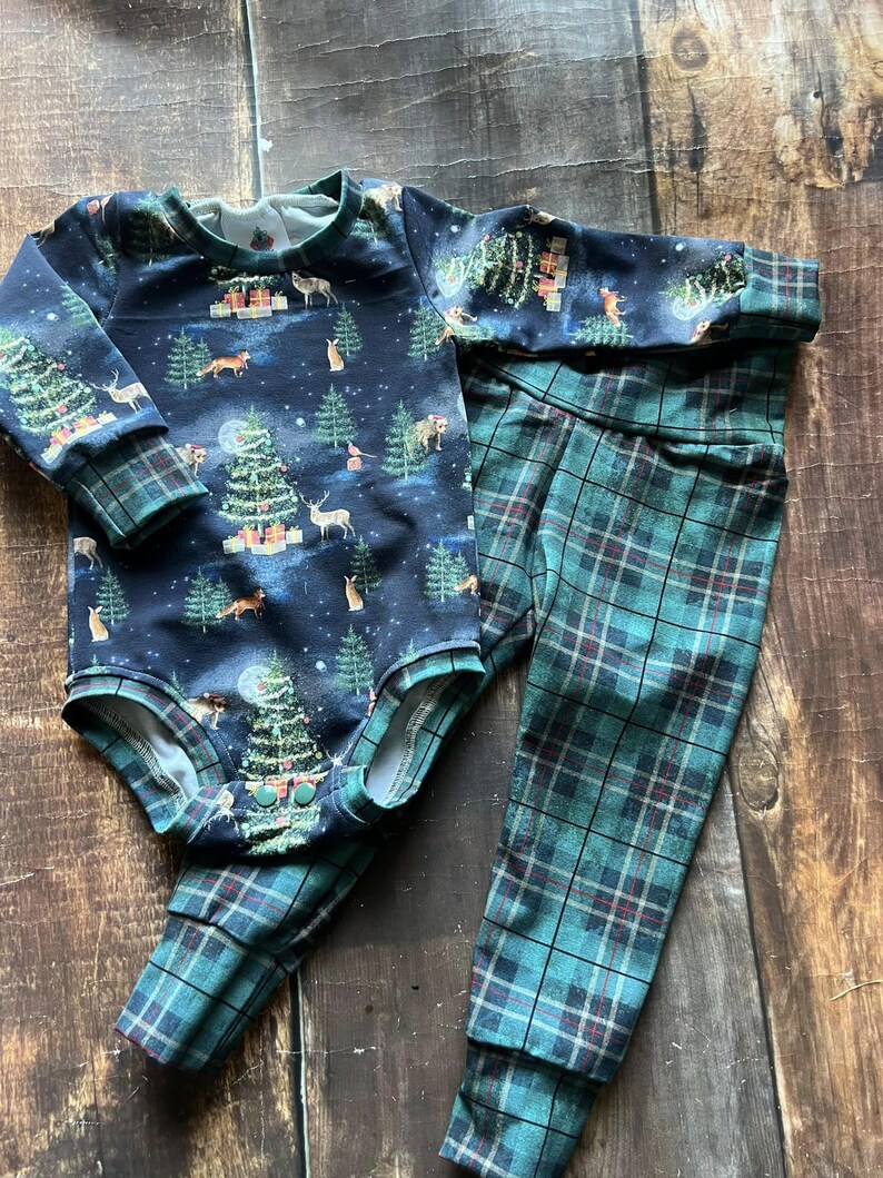 Op de afbeelding: Een baby-outfitset met een longsleeve bodysuit met een donkerblauwe achtergrond en een feestelijke kerstboomprint. De broek is groen en zwart geruit.