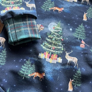Op de afbeelding: Zwarte stof met een herhalend patroon van kerstbomen, herten, vossen, konijnen en vogels. De stof heeft een donkerblauwe achtergrond met witte sterren.