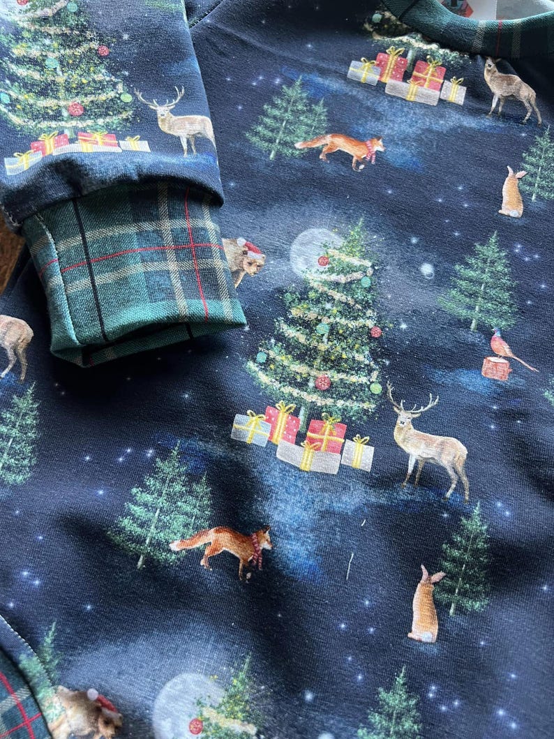 Op de afbeelding: Zwarte stof met een herhalend patroon van kerstbomen, herten, vossen, konijnen en vogels. De stof heeft een donkerblauwe achtergrond met witte sterren.