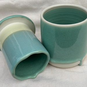 May include: A two-piece ceramic butter keeper set featuring a cylindrical base and a matching lid. The exterior is finished in a glossy teal glaze that drips towards the base, while the interior and upper rim reveal a smooth, off-white ceramic surface.