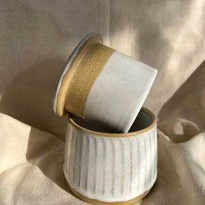 May include: A two-piece ceramic butter keeper featuring a speckled tan base and a white glazed top. The bottom container has vertical ribbed detailing, while the lid has a smooth finish. The pieces are stacked with the lid resting at an angle.