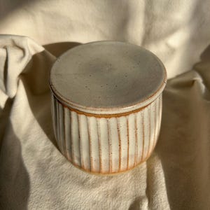 May include: A cylindrical ceramic jar with a matching lid, featuring a cream-coloured glaze with subtle brown speckles. The exterior surface is decorated with vertical fluted grooves, and the base and rim show exposed reddish-brown clay.