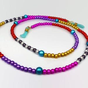 May include: A multi-colored beaded eyeglass chain featuring a repeating pattern of metallic gold, silver, red, purple, magenta, and bright blue spherical beads. The ends of the chain are finished with light blue silicone loops for attachment.