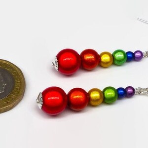 May include: Rainbow colored beaded dangle earrings with silver hooks. The beads are red, orange, yellow, green, blue, and purple. The earrings are about the size of a US one pound coin.
