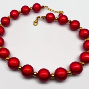 Può includere: Una collana di perle rosse con accenti dorati. Le perle sono grandi e rotonde, e la collana è infilata su una catena dorata.