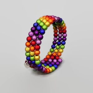 Puede incluir: Un brazalete de cuentas de colores arcoíris con un dije de corazón plateado. El brazalete está hecho de pequeñas cuentas redondas en tonos de rojo, naranja, amarillo, verde, azul, índigo y violeta.
