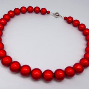 Peut inclure: Un collier composé de perles sphériques rouge vif et brillantes dont la taille augmente progressivement vers le centre. Les perles sont enfilées ensemble et fixées par un fermoir magnétique sphérique de couleur argentée à l'arrière du collier.