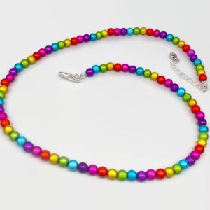 Puede incluir: Un collar de cuentas de colores con un arcoíris de colores, incluyendo rojo, naranja, amarillo, verde, azul y morado. El collar tiene un cierre plateado y una cadena extensora. Las cuentas tienen aproximadamente 0,6 cm de diámetro.