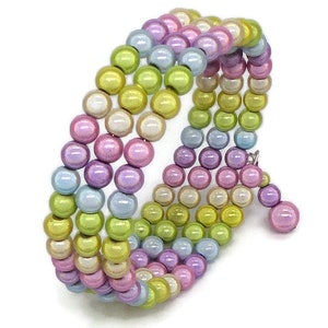 Può includere: Un bracciale avvolgente a più fili con file di perline sferiche iridescenti in tonalità pastello di verde lime, lavanda e azzurro. Il bracciale ha un design a spirale flessibile e include un piccolo ciondolo composto da due perline.