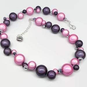 Op de afbeelding: Een ketting met een mix van roze en donkerpaarse kralen. De kralen variëren in grootte en zijn geregen met een zilverkleurige ketting en sluiting. De ketting heeft een hartvormige bedel.