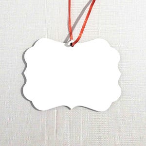 May include: White ceramic ornament with a scalloped edge and a red ribbon attached. The ornament is blank and ready to be personalized.