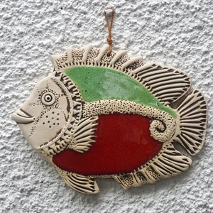 Op de afbeelding: Een keramische vis wanddecoratie met een rode, groene en witte glazuur. De vis heeft een gedetailleerd ontwerp met een textuur.