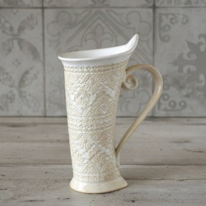 Peut inclure: Une tasse en céramique blanche avec un motif en relief complexe. La tasse a une poignée incurvée unique et une forme haute et mince.