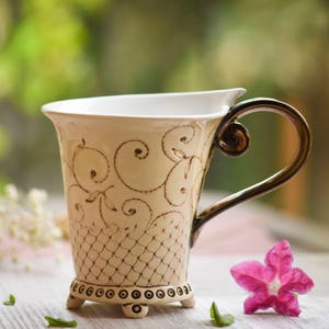 Peut inclure: Une tasse en céramique blanche avec une anse brune et un motif floral complexe. La tasse a une base décorative avec de petits pieds et est posée sur une surface en bois clair.