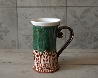 Könnte beinhalten: Eine grüne Keramiktasse mit braunem Henkel und weißen geometrischen Mustern am Boden. Die Tasse hat einen weißen Rand und steht auf einer hellbraunen Holzoberfläche.