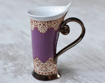 May include: A purple ceramic mug with a white interior and a gold-colored handle. The mug is decorated with a lace pattern around the top and bottom.