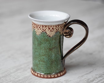 May include: A green ceramic mug with a brown handle and a gold and brown lace design around the rim.